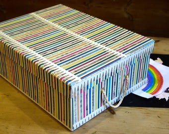May include: A rectangular storage box with a lid, featuring a repeating pattern of multicolored pencils arranged vertically. The box includes a single white rope handle on the front side, and the exterior surface is covered in a vibrant pencil print.