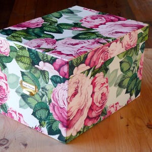 May include: A white storage box with a floral pattern featuring pink roses and green leaves. The box has a gold metal label holder.