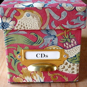 Puede incluir: Caja decorativa con tapa, adornada con un diseño floral y de pájaros en rojo, verde y dorado. Tiene un portaetiquetas dorado con la palabra "CDs". La caja está diseñada para el almacenamiento.