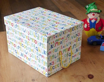 May include: A white storage box with a colorful alphabet pattern and yellow rope handles. The box is sitting on a wooden surface.