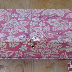 May include: A pink box with a white and brown floral pattern. The box has a ribbon tied around it and a metal eyelet on the top.