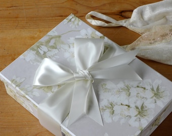 Puede incluir: Una caja de regalo blanca con un diseño floral y un gran lazo de satén blanco. La caja está sobre una superficie de madera.