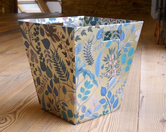 May include: A decorative storage basket with a floral pattern in shades of blue, green, and gold. The basket is made of cardboard and has a square shape.