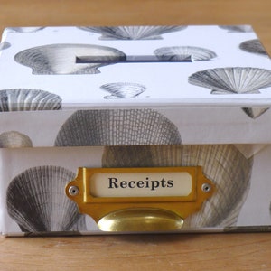 May include: A white box with a seashell pattern and a gold label that says "Receipts". The box has a gold handle and a slot on the top.