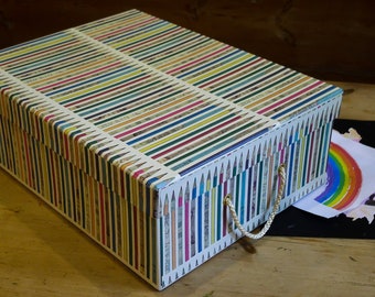 May include: A white box with a colorful pencil pattern and rope handles. The box is sitting on a wooden surface with a drawing of a rainbow on a piece of paper next to it.
