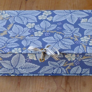 Puede incluir: Una caja azul con un patrón floral blanco y azul y una cinta blanca atada alrededor de la tapa. La caja tiene un cierre de latón.