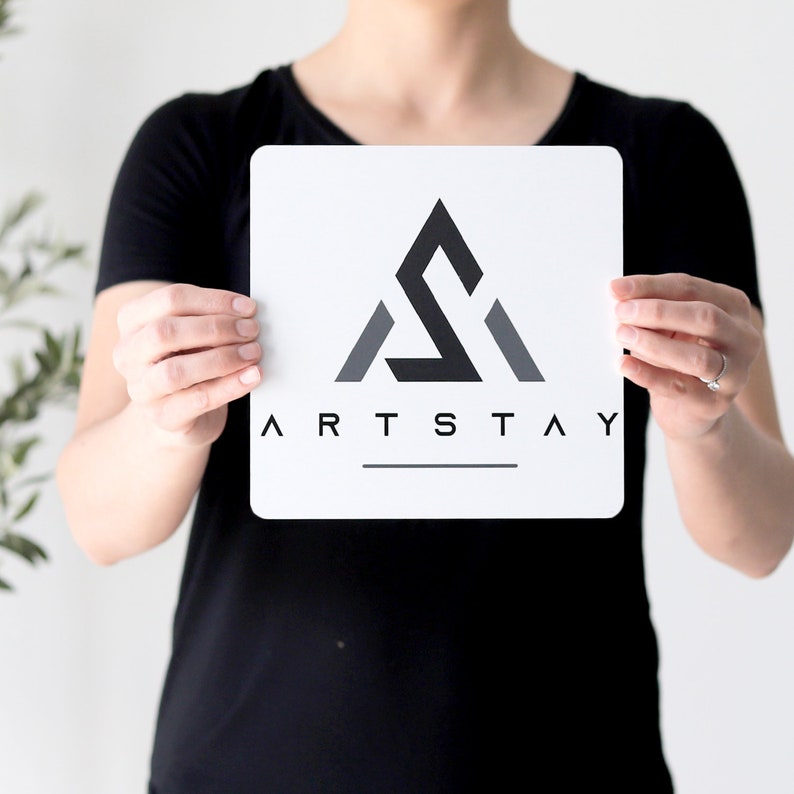 Op de afbeelding: Een wit bord met een zwart en grijs geometrisch logo met de tekst "ARTSTAY" in zwarte letters.