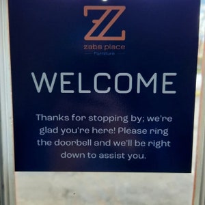 Op de afbeelding: Een blauw bord met witte tekst dat zegt: "Z zabs place Furniture WELCOME Thanks for stopping by; we're glad you're here! Please ring the doorbell and we'll be right down to assist you."