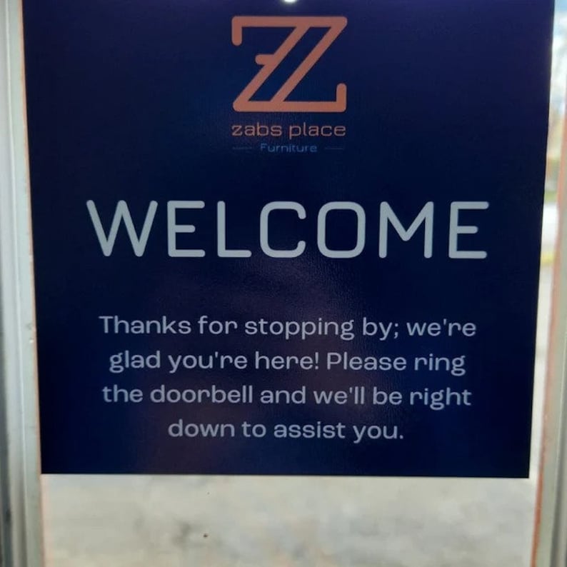 Op de afbeelding: Een blauw bord met witte tekst dat zegt: "Z zabs place Furniture WELCOME Thanks for stopping by; we're glad you're here! Please ring the doorbell and we'll be right down to assist you."