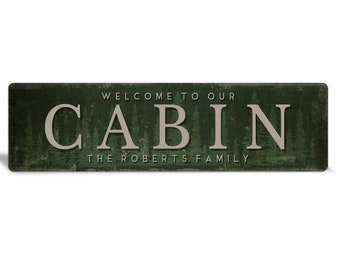 May include: A green wooden sign with the text "Welcome to our Cabin The Roberts Family" in white lettering. The sign has a rustic, distressed look and features a forest background.