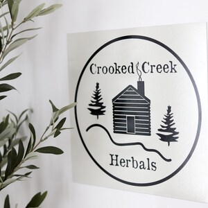 Op de afbeelding: Zwart-wit logo met de tekst "Crooked Creek Herbals" in een cirkel. Het logo toont een hut met een schoorsteen, twee dennenbomen en een gebogen lijn die een heuvel voorstelt.
