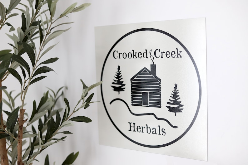 Op de afbeelding: Zwart-wit logo met de tekst "Crooked Creek Herbals" in een cirkel. Het logo toont een hut met een schoorsteen, twee dennenbomen en een gebogen lijn die een heuvel voorstelt.