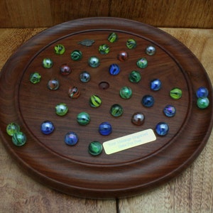 May include: A wooden marble game board with a circular playing surface and a personalized plaque that reads "Your Name Engraved Here". The board is surrounded by a dark brown wooden frame. There are 28 marbles on the board, in various colors, including green, blue, and red.