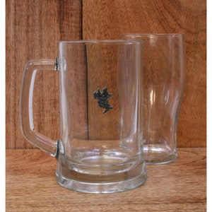 May include: Two clear glass beer mugs with a handle and a decorative emblem. The mugs are empty and sit on a wooden surface. One mug has a handle, and the other is a pint glass.