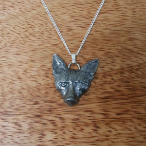 May include: A silver-colored pendant necklace featuring a detailed fox head. The fox pendant is attached to a delicate silver chain. The necklace is displayed against a wooden background.