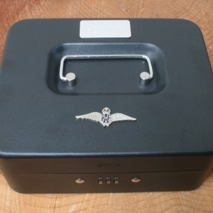 Puede incluir: Una caja de metal negra con una cerradura de combinación y una manija plateada. La caja tiene un emblema plateado de la Royal Air Force en la parte superior.