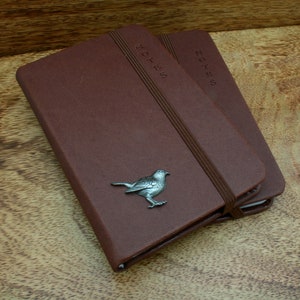 May include: Two brown leather bound notebooks with the word "NOTES" embossed on the front. The top notebook has a silver bird charm attached.