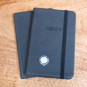 May include: Two gray hardcover notebooks with black elastic closures. The notebook on top has the word "notes" embossed on the front cover. The notebook on the bottom has a small round pin with a blue and white design.
