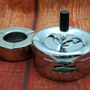 May include: A chrome ashtray with a rotating lid and a fish design. The ashtray has a separate, smaller ashtray for cigarette butts.