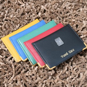 May include: Five leather travel card holders in different colors: yellow, blue, green, red, and black. The black card holder has gold trim and the text "Travel Card" embossed on the front.