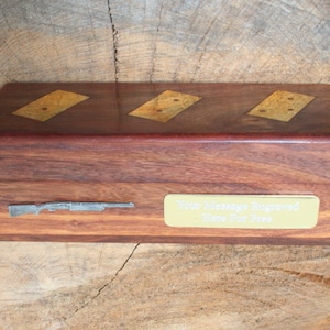 Puede incluir: Una caja de madera con una placa de metal dorado grabada con el texto "Your Message Engraved Here For Free". La caja tiene tres dominós de oro incrustados y un diseño de escopeta de plata.