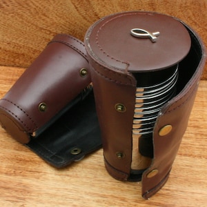 May include: A brown leather case with a silver metal object inside. The case is cylindrical with a snap closure and a small silver fish emblem on the top. The case is open, revealing the contents.