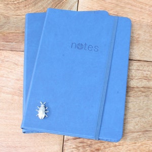 May include: Two blue notebooks with a silver bug charm on the front. The notebook on top has the word "notes" printed on the front.