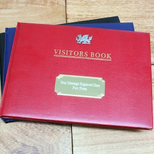 May include: Red leather visitors book with a gold embossed dragon and the text "VISITORS BOOK". A gold plate with the text "Your Message Engraved Here For Free" is on the front cover.