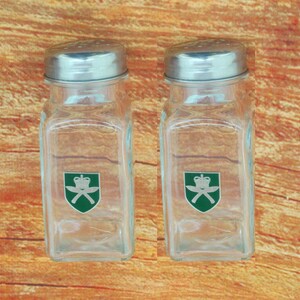 May include: Two clear glass salt and pepper shakers with silver lids. Each shaker has a green and silver emblem with a crown and crossed swords.