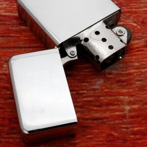 Puede incluir: Un encendedor plateado con una tapa abatible abierta, que revela la piedra y la mecha en el interior. El encendedor est&aacute; sobre una superficie con un patr&oacute;n rojo y naranja.