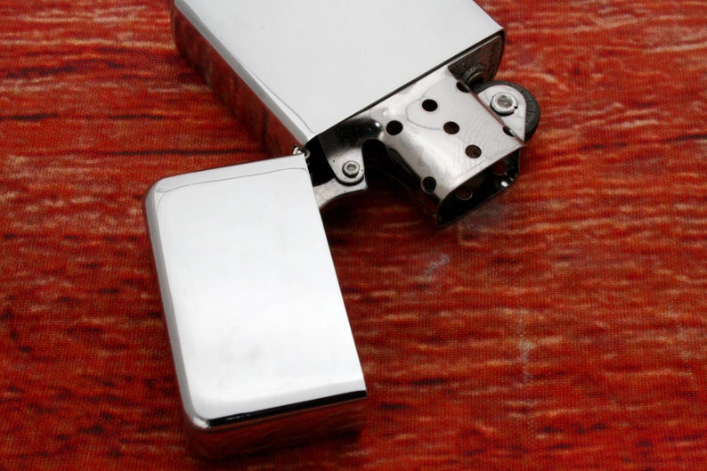 Puede incluir: Un encendedor plateado con una tapa abatible abierta, que revela la piedra y la mecha en el interior. El encendedor est&aacute; sobre una superficie con un patr&oacute;n rojo y naranja.