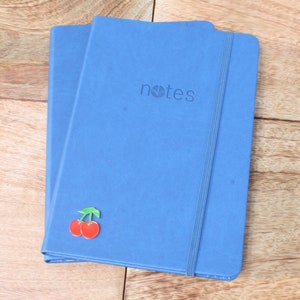 May include: Two blue notebooks with a cherry sticker on the front. The notebook on top has the word "notes" printed on the front.