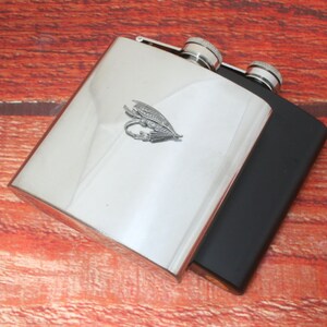 May include: A silver and black stainless steel hip flask with a fly fishing lure design. The flask has a screw-on cap.