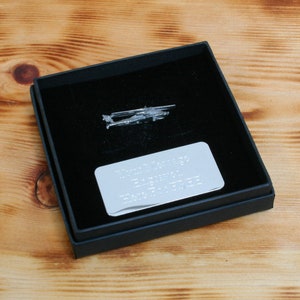 Puede incluir: Un pin de helicóptero plateado en una caja de regalo negra con una placa grabada plateada. La placa tiene texto que es difícil de leer. La caja está sobre una superficie de madera.