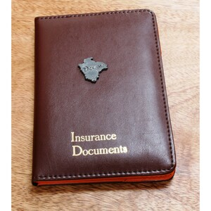 Pode incluir: Um porta-documentos de seguro de couro marrom com uma inscrição dourada que diz "Insurance Documents". O porta-documentos tem um pequeno emblema de metal do estado do Texas.