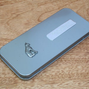 May include: A silver metal tin with a cat figurine on the front. The tin has a silver label with the text "Designed in England" on it.