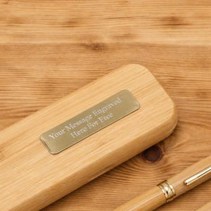 K&ouml;nnte beinhalten: Ein helles Bambus-Stift- und Etui-Set. Das Etui hat ein rechteckiges Metallschild mit dem Text "Your Message Engraved Here For Free". Der Stift hat goldfarbene Akzente.
