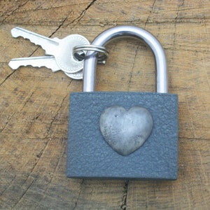 Puede incluir: Un candado gris con un diseño en forma de corazón en la parte delantera. El candado está unido a dos llaves.