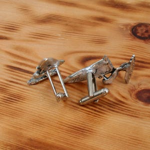 May include: Two silver cufflinks with detailed designs. One cufflink features a bird design, while the other features a geometric design.