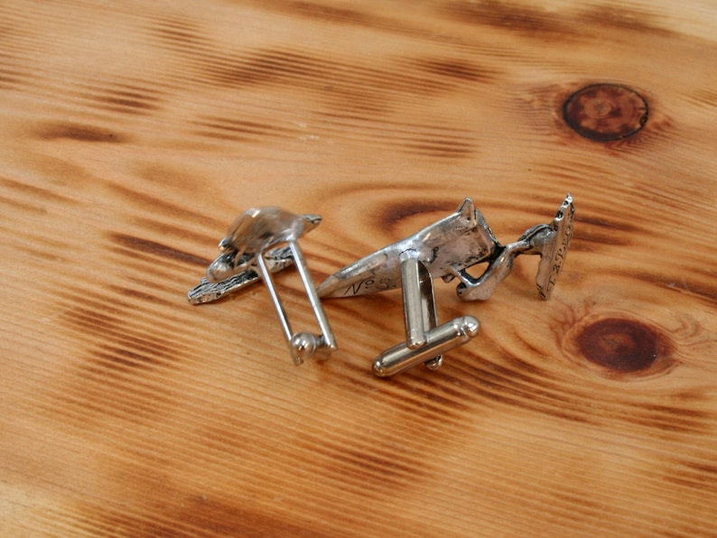May include: Two silver cufflinks with detailed designs. One cufflink features a bird design, while the other features a geometric design.