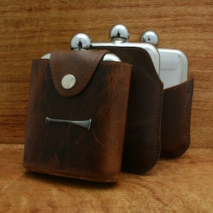 May include: Three stainless steel hip flasks with brown leather cases. The flask on the left has a small metal design on the front.