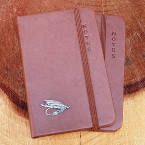May include: Two brown leather bound notebooks with the word "NOTES" embossed on the front cover. The notebook on the left has a silver fly fishing lure attached to the front cover.