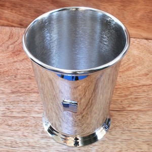 Könnte beinhalten: Eine silberne Metalltasse mit einer kleinen amerikanischen Flagge an der Seite. Die Tasse steht auf einer Holzoberfläche.