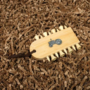 May include: A wooden golf tee holder with a metal tractor emblem. The holder is light brown and has several golf tees attached. A dark brown leather strap is attached to the holder.