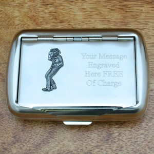 Può includere: Un porta sigarette in metallo argentato con un coperchio incernierato. La custodia ha una piccola immagine incisa di una persona sul davanti. Il testo "Your Message Engraved Here FREE Of Charge" è inciso sul coperchio.