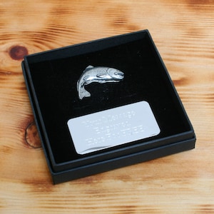 Pode incluir: Uma figura de truta prateada em uma caixa de veludo preto com uma placa prateada gravada com uma mensagem personalizada.