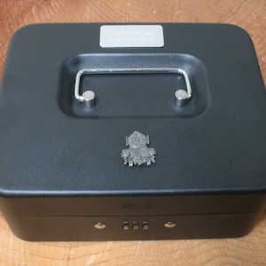 Puede incluir: Caja de metal negra con una cerradura de combinación y una manija plateada. La caja tiene una placa plateada con el texto "Cornish Steam Railway" y un emblema plateado de un tren de vapor.