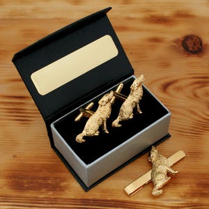 May include: Gold-coloured wolf cufflinks and tie clip set. The cufflinks feature detailed wolf figures, presented in a black and silver gift box with a gold nameplate. The tie clip has a single wolf design.