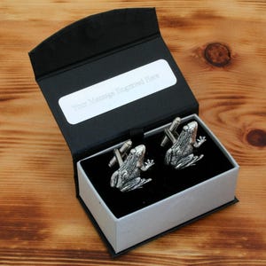 May include: A black gift box containing a silver frog cufflink set. The box has a white label with the text "Your message engraved here".
