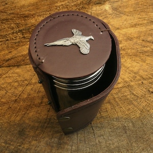 Puede incluir: Un recipiente de cuero marrón con un emblema de faisán plateado en la tapa. El recipiente está abierto, revelando el contenido en el interior. El cuero tiene un diseño cosido y está asegurado con remaches metálicos.